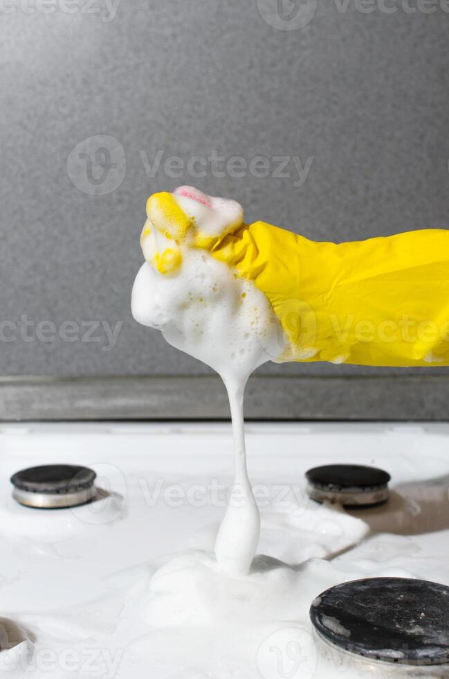 Housewife in glove scrubbing the surface of gas stove with sponge and chemical foam. Vertical photo. Home cleaning concept, kitchen hygiene photo