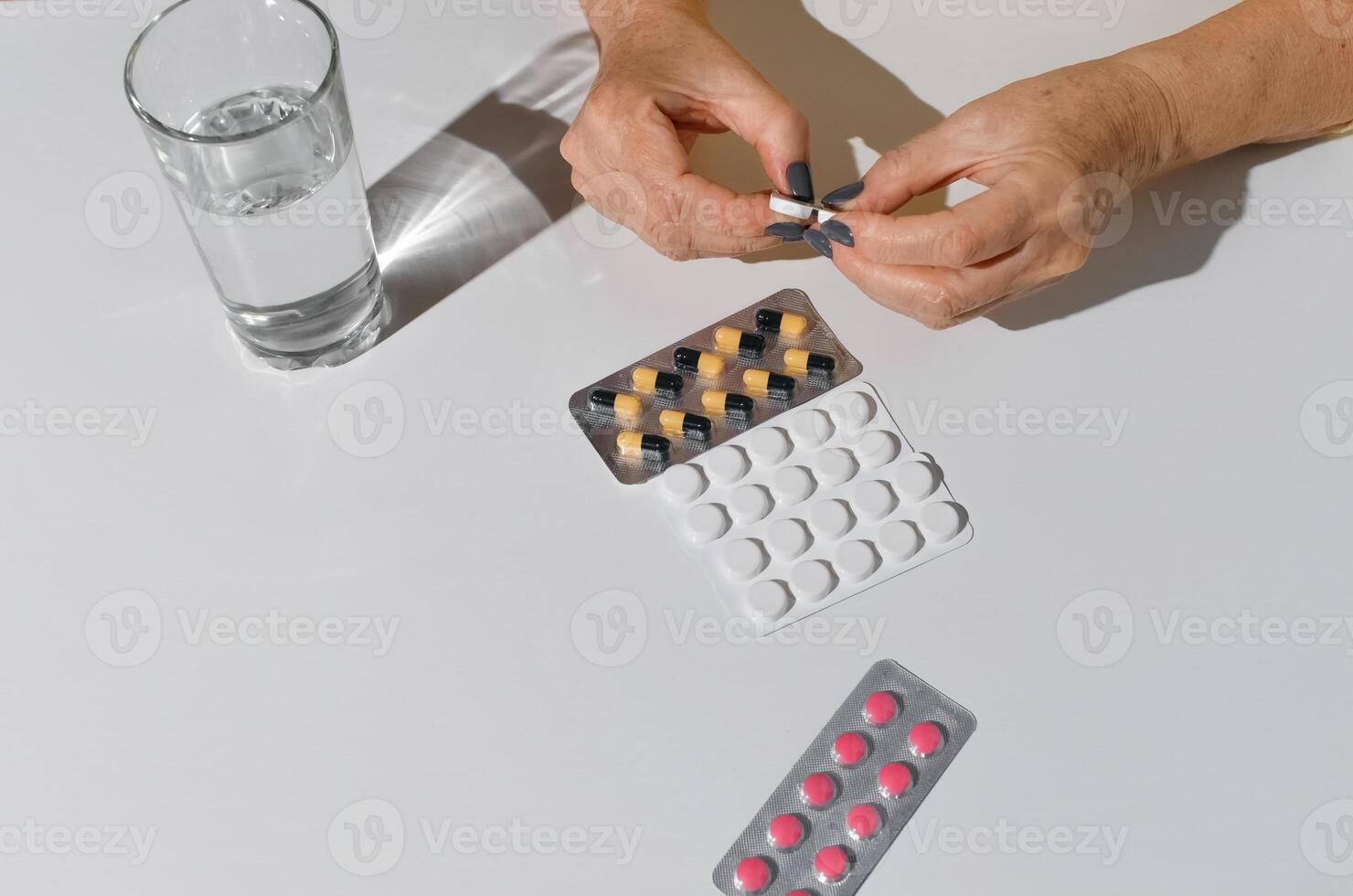 Elderly woman's hands opening package of medicines. Tablets and glass of water on white table, top view. photo