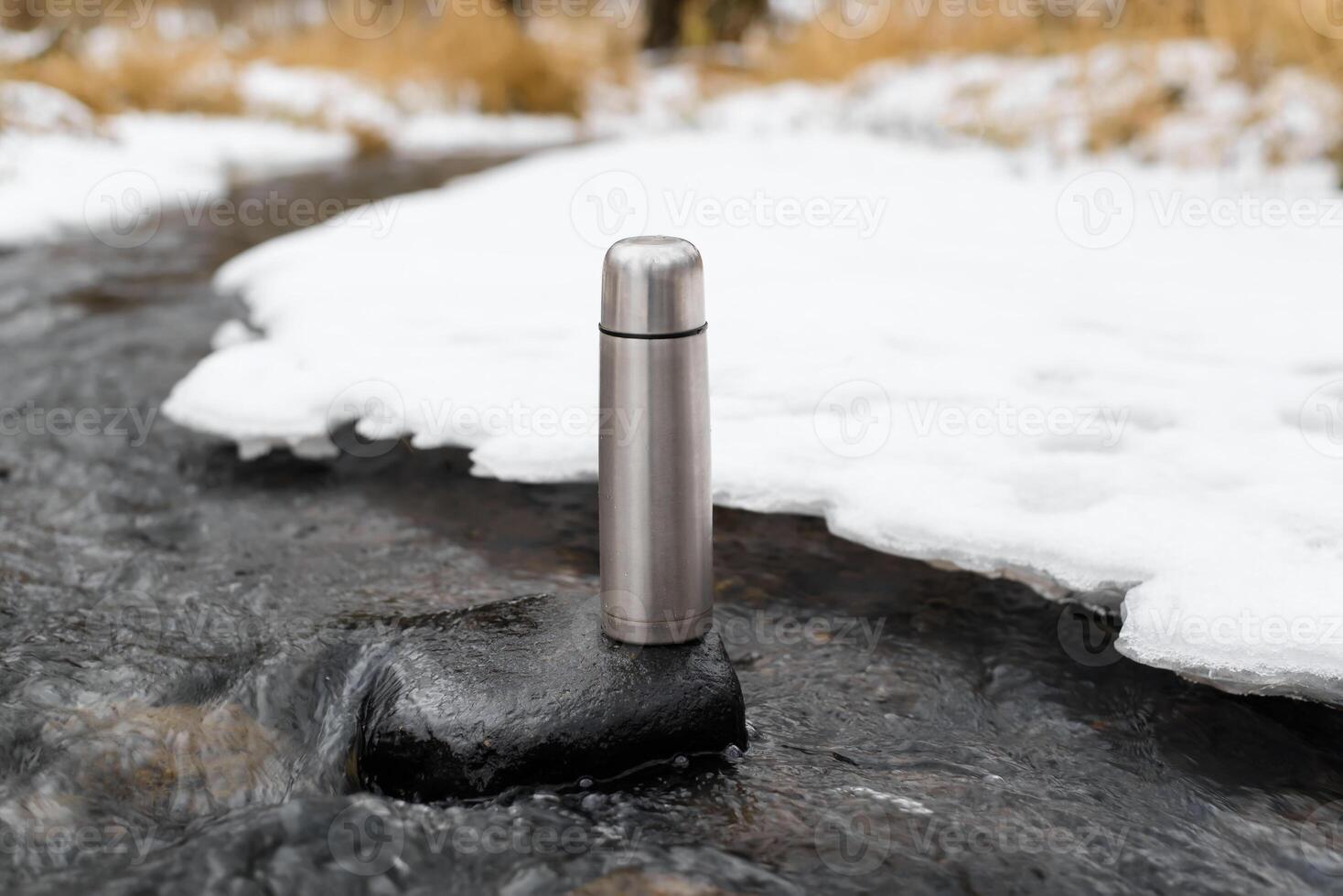 Steel vacuum thermos in a winter stream. Hiking flask standing on a stone outside. Adventure, hike, travel concept. photo