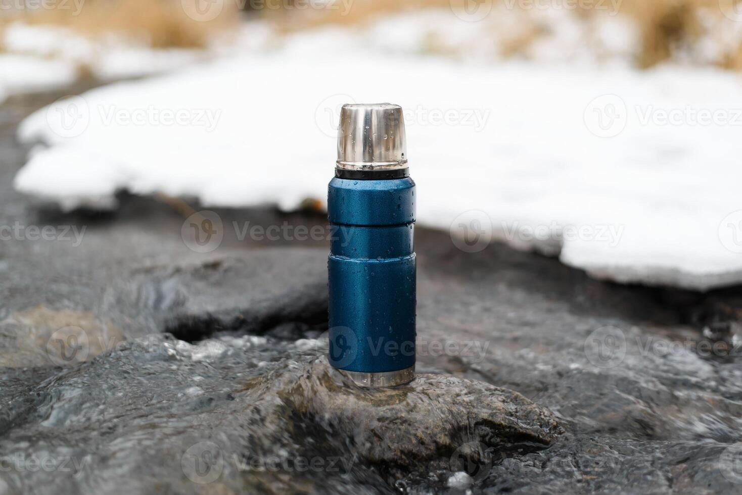 Blue vacuum thermos close-up outside in winter. Camping thermos standing on a stone in a winter stream. Adventure concept, winter hike, travel. photo