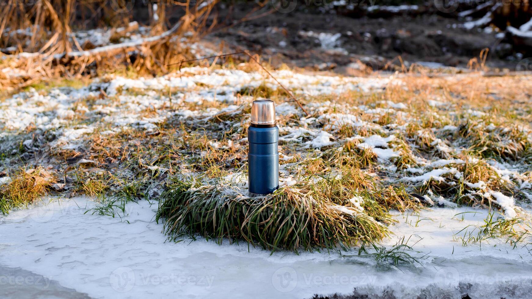 Thermos standing outside in the winter forest at dawn. Vacuum blue hiking flask. Travel concept, adventure, winter warming drink. photo