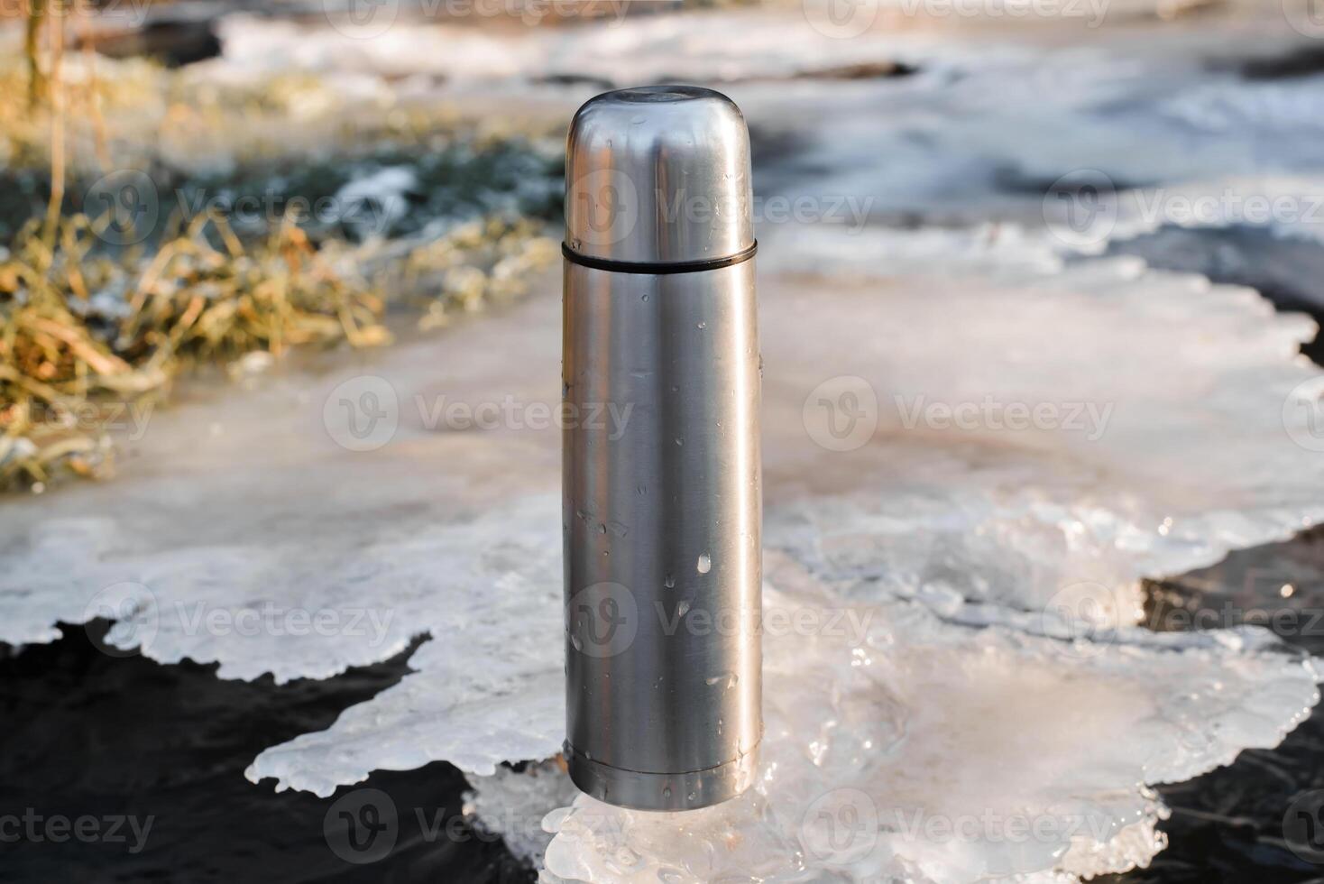 Thermos standing on ice, close-up. Vacuum steel flask for hiking. Travel concept, warming drink in winter. photo