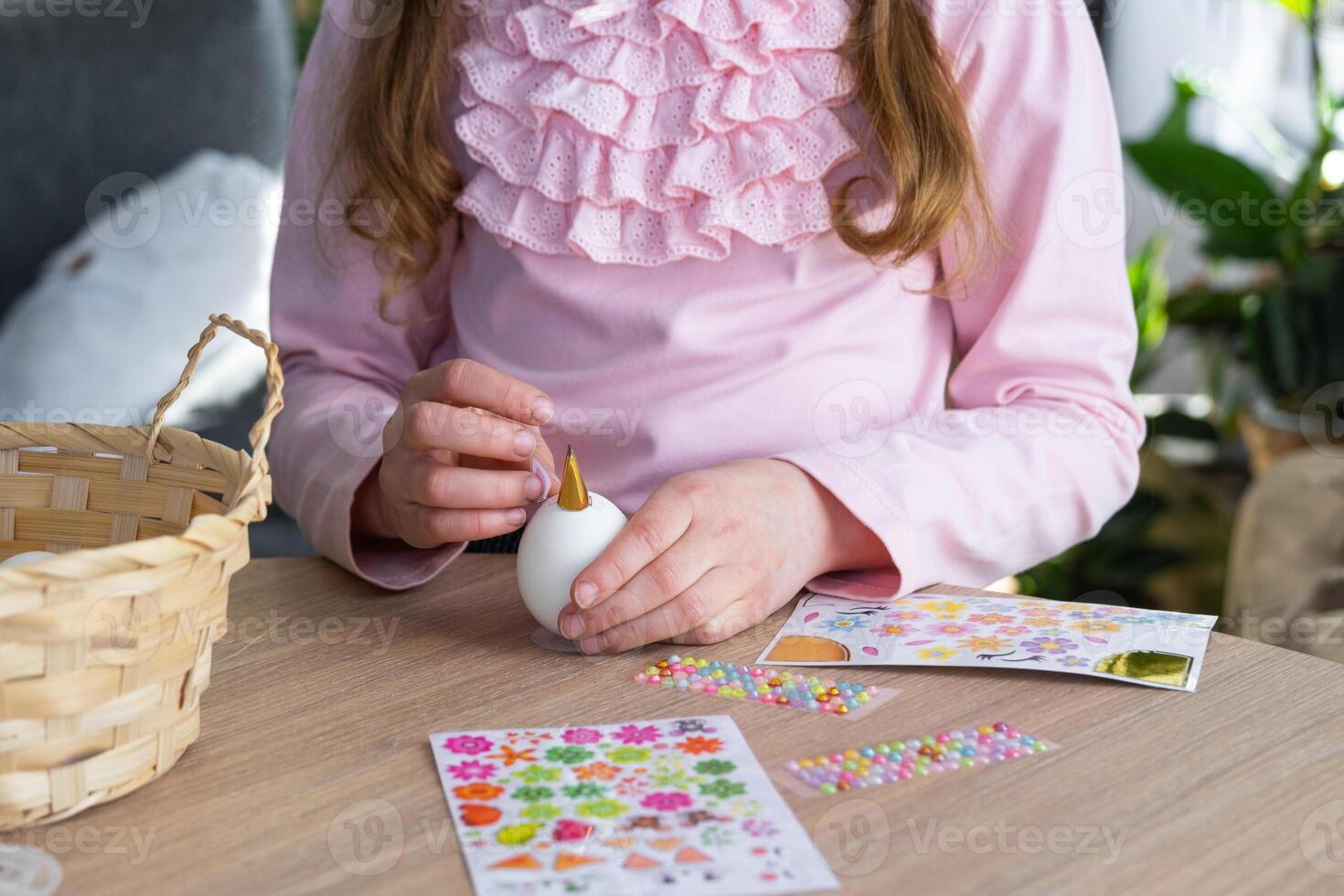A cute girl with pink bunny ears makes an Easter craft - decorates an egg in the form of a unicorn with rhinestones, horn, flowers in the interior of a house with plants. photo