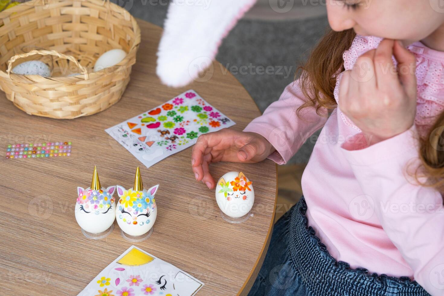 A cute girl with pink bunny ears makes an Easter craft - decorates an egg in the form of a unicorn with rhinestones, horn, flowers in the interior of a house with plants. photo