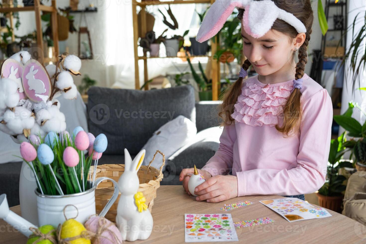 A cute girl with pink bunny ears makes an Easter craft - decorates an egg in the form of a unicorn with rhinestones, horn, flowers in the interior of a house with plants. photo