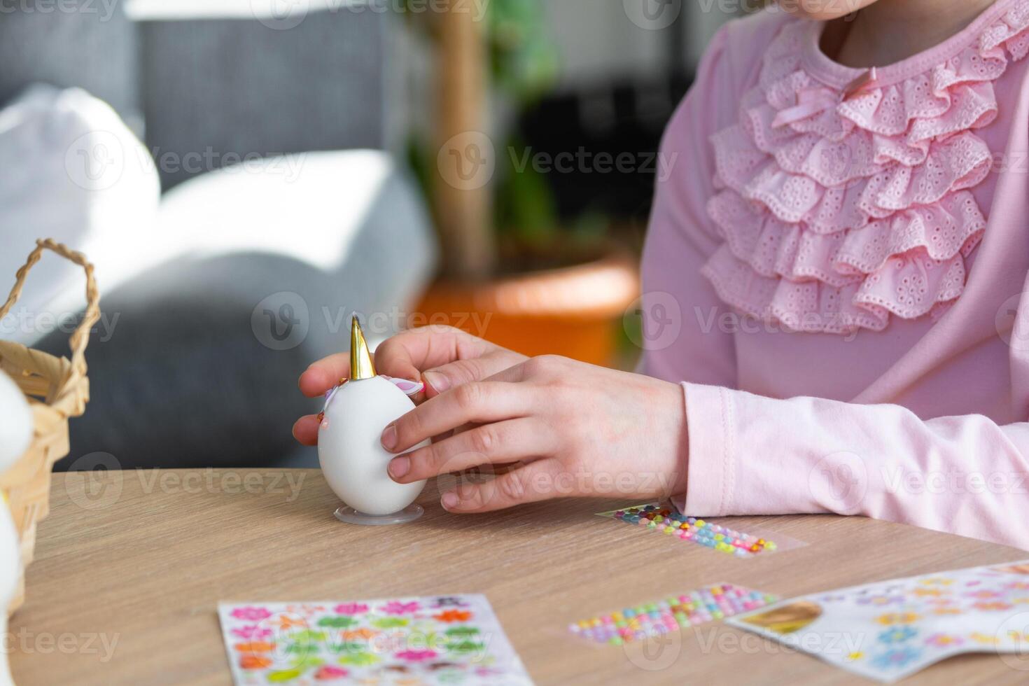 A cute girl with pink bunny ears makes an Easter craft - decorates an egg in the form of a unicorn with rhinestones, horn, flowers in the interior of a house with plants. photo