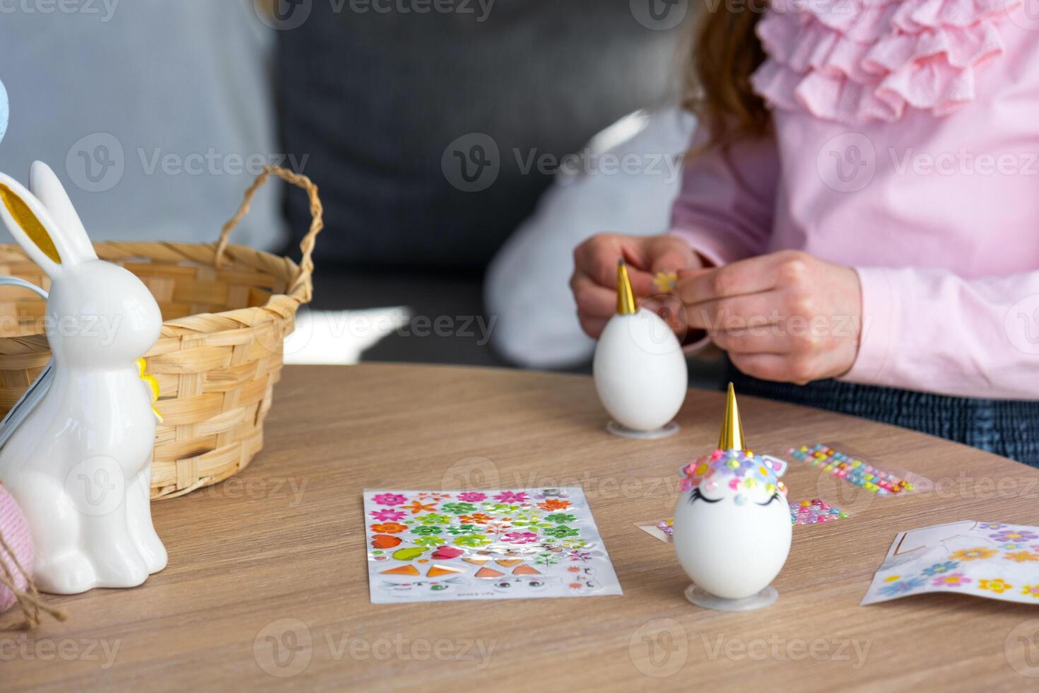 A cute girl with pink bunny ears makes an Easter craft - decorates an egg in the form of a unicorn with rhinestones, horn, flowers in the interior of a house with plants. photo