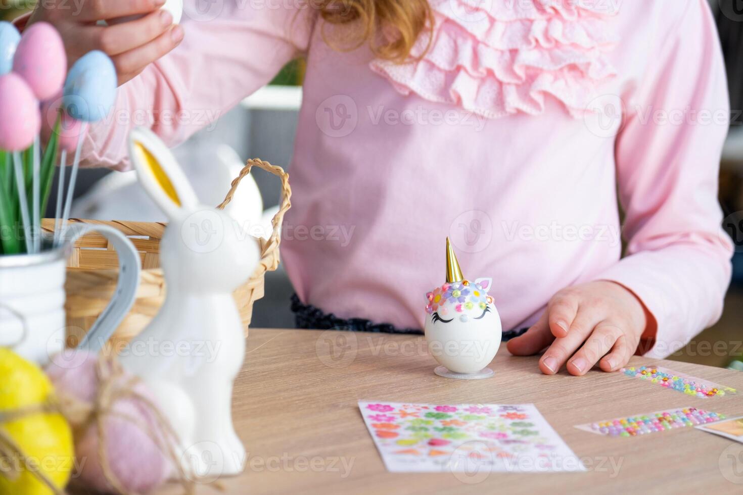 A cute girl with pink bunny ears makes an Easter craft - decorates an egg in the form of a unicorn with rhinestones, horn, flowers in the interior of a house with plants. photo