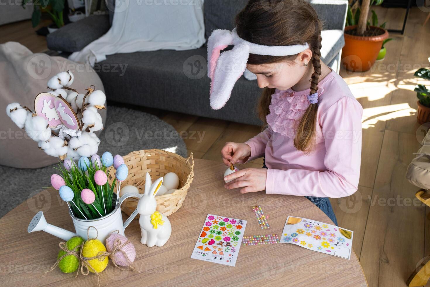 A cute girl with pink bunny ears makes an Easter craft - decorates an egg in the form of a unicorn with rhinestones, horn, flowers in the interior of a house with plants. photo