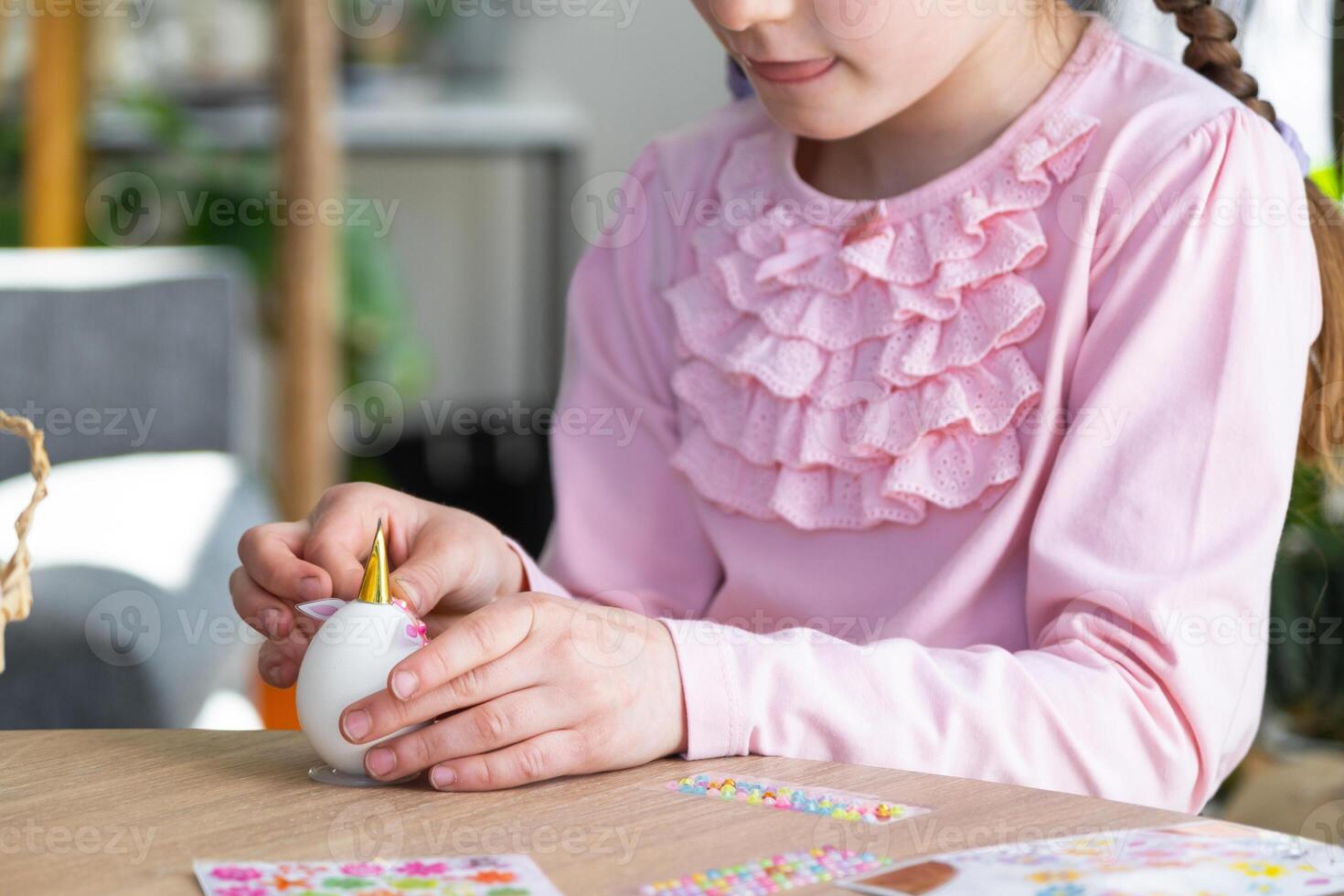 A cute girl with pink bunny ears makes an Easter craft - decorates an egg in the form of a unicorn with rhinestones, horn, flowers in the interior of a house with plants. photo