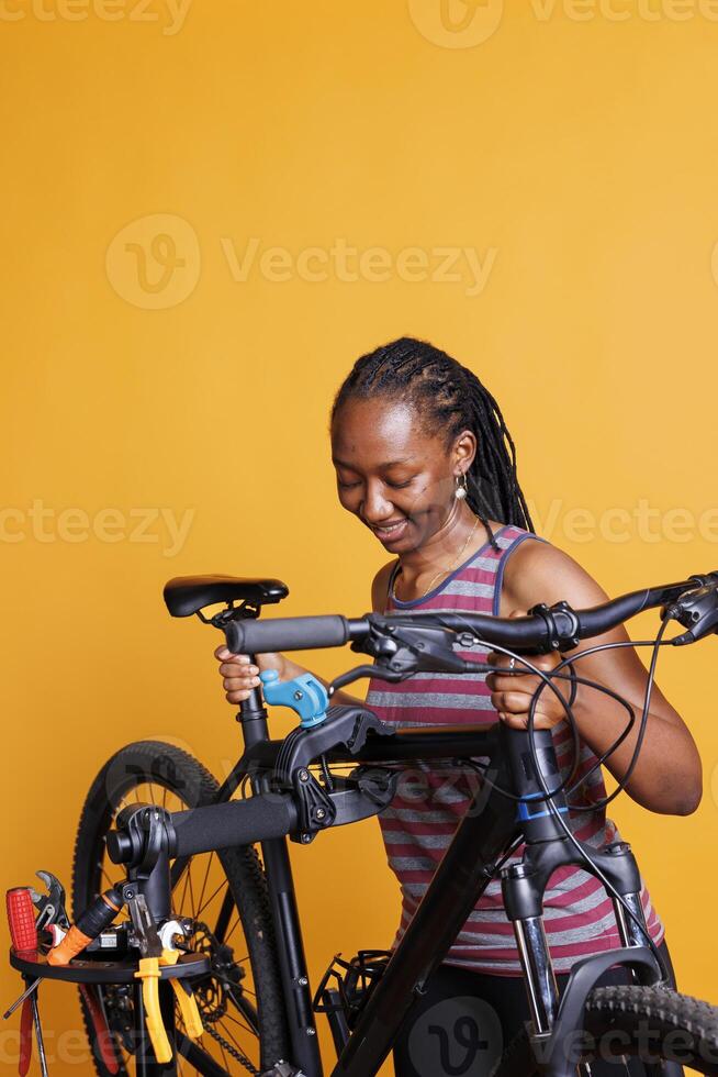 Active healthy female securing bike on repair-stand for repairing against yellow background. Image showing sporty black woman adjusting bicycle height, ready for yearly maintenance. photo