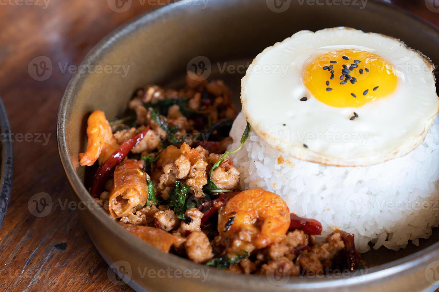 Asian Thai food mouth watering dish of spicy basil shrimp and pork over rice topping with a perfectly fried egg and black sesame seeds served in a rustic bowl on wooden table photo