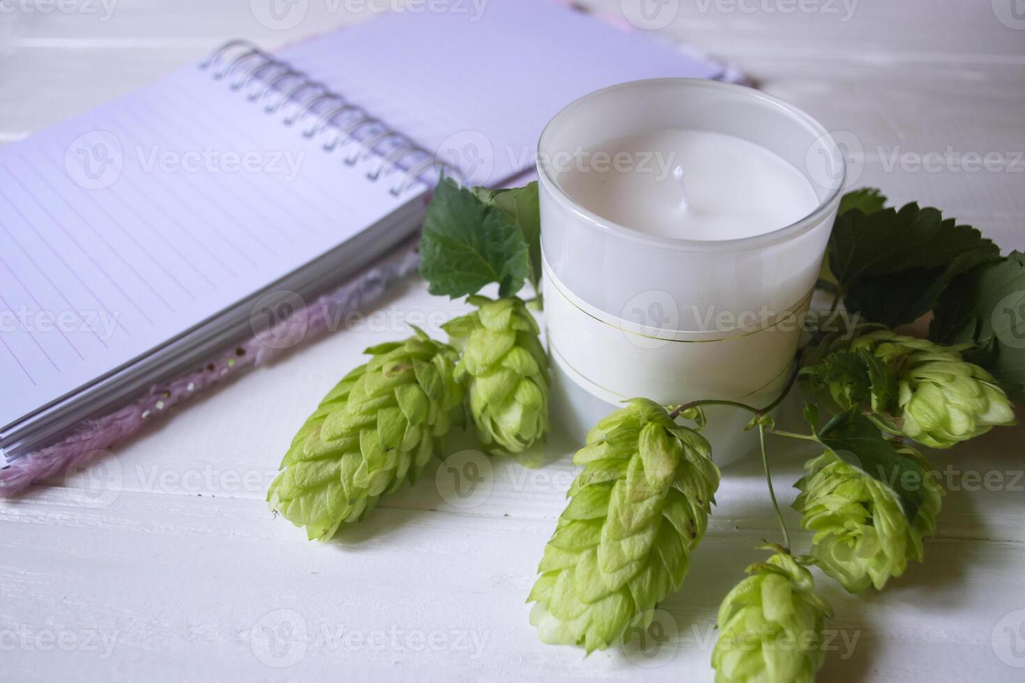 The opened notepad, pen, white candle, glasses and branches of hops as decoration on a white wooden table. Desktop still life with space for text. photo