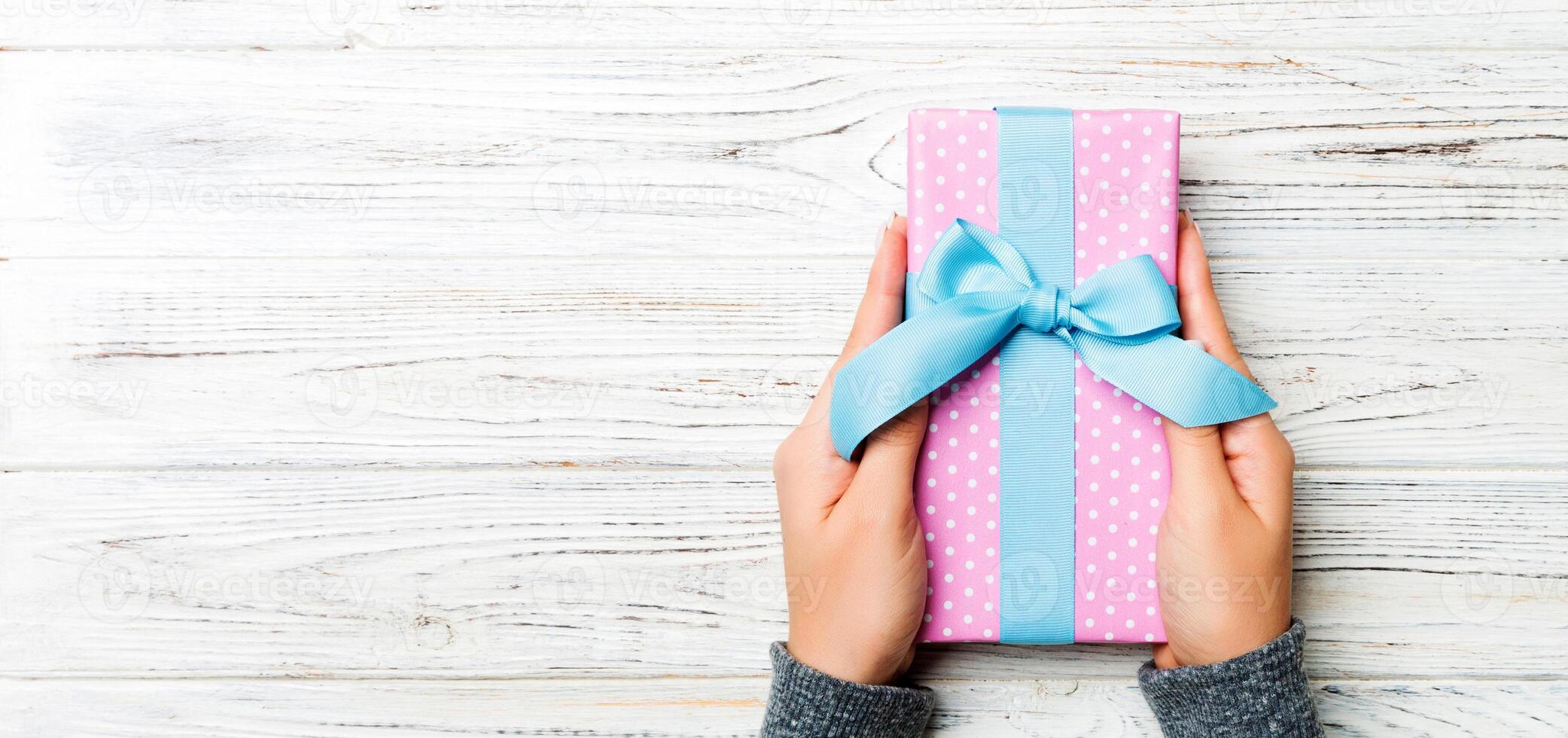 Woman hands give wrapped christmas or other holiday handmade present in colored paper . Present box, decoration of gift on white rustic wooden table, top view with copy space photo
