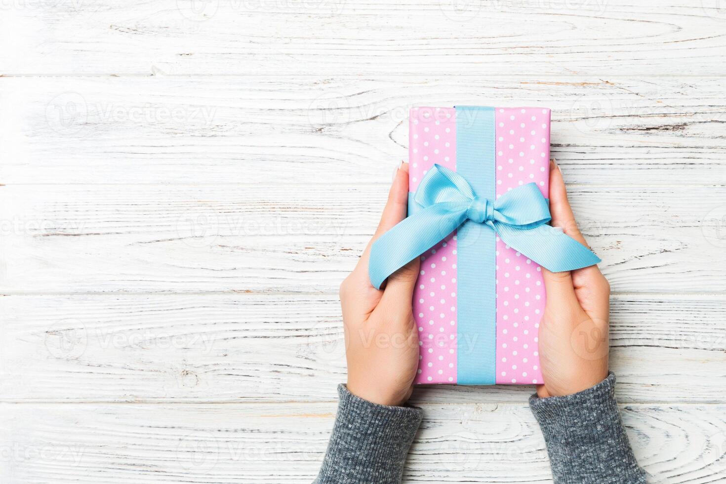 Woman hands give wrapped christmas or other holiday handmade present in colored paper . Present box, decoration of gift on white rustic wooden table, top view with copy space photo