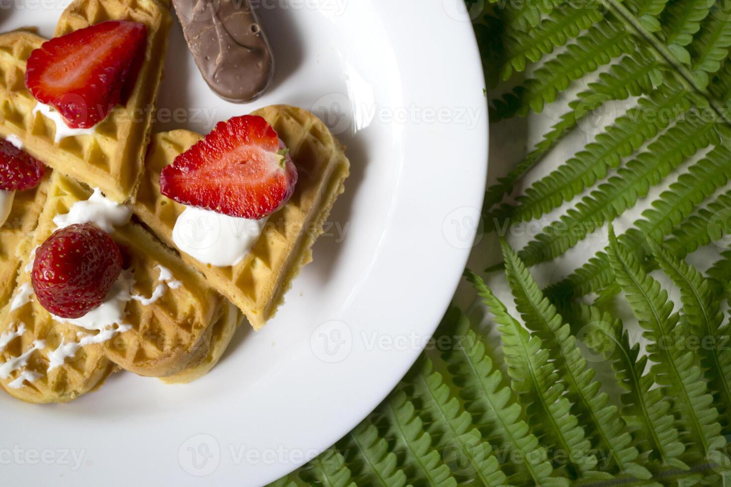Waffles with strawberry on a plate, green leaves of fern on a table. Close up. Beautiful and tasty breakfast. photo