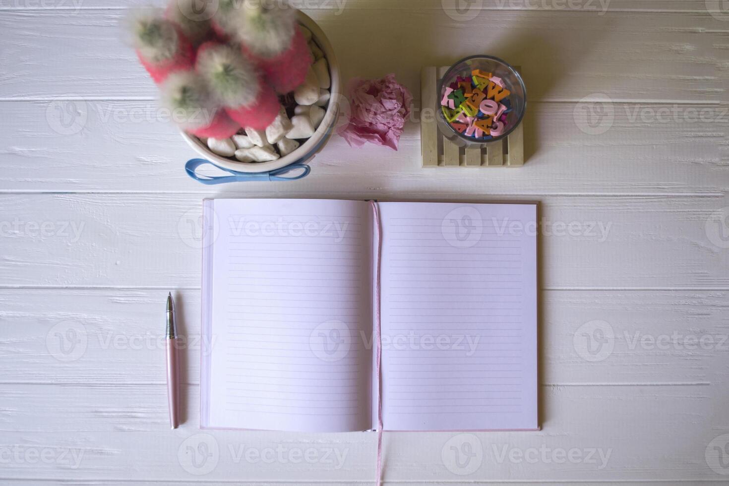 A notepad with pen, pink cactus, crumpled sheet of paper and multicolored letters on a white wooden desktop. photo