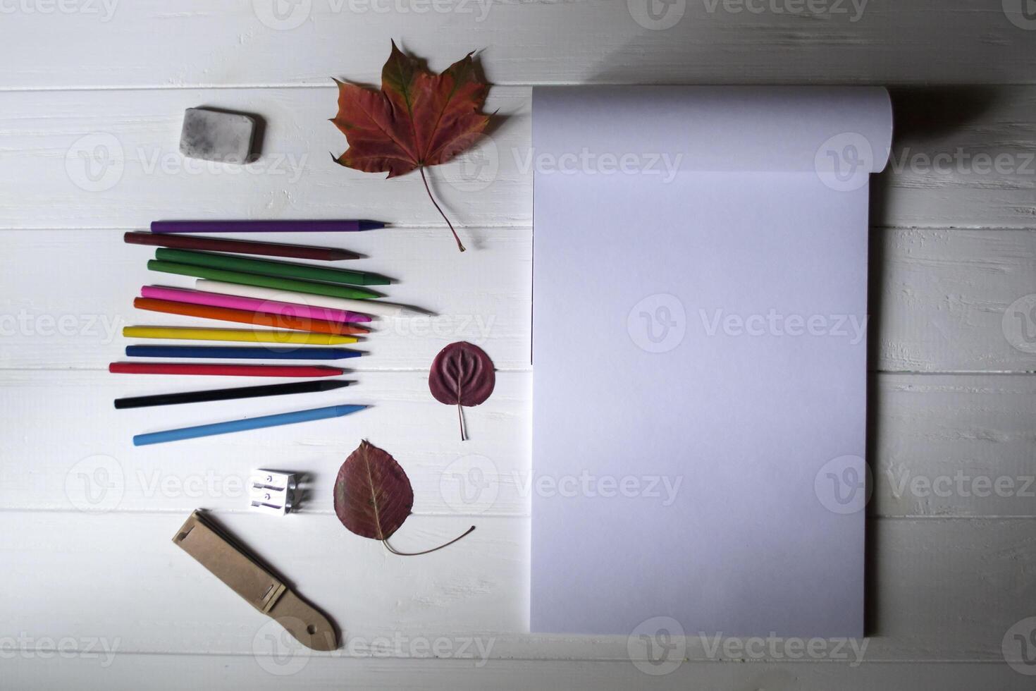 Empty sketch pad, colorful pencils, other equipment for drawing and fallen leaf on the white wooden desktop. Art background with place for text. photo