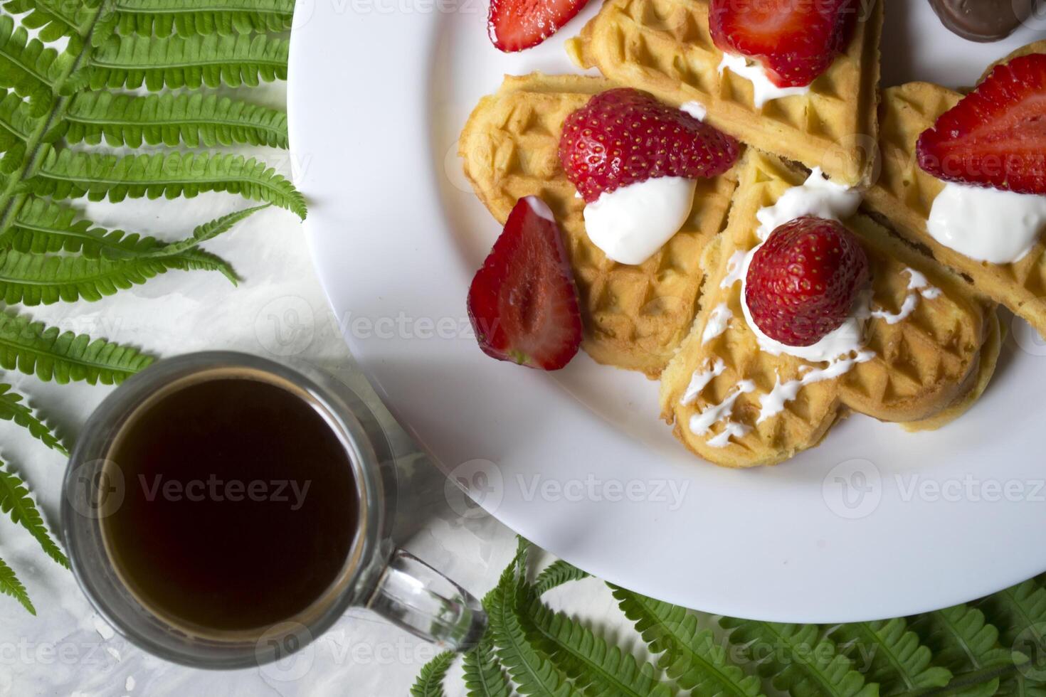 Waffles with strawberry, a cup of coffee and green leaves of fern on a table. Beautiful and tasty breakfast. photo