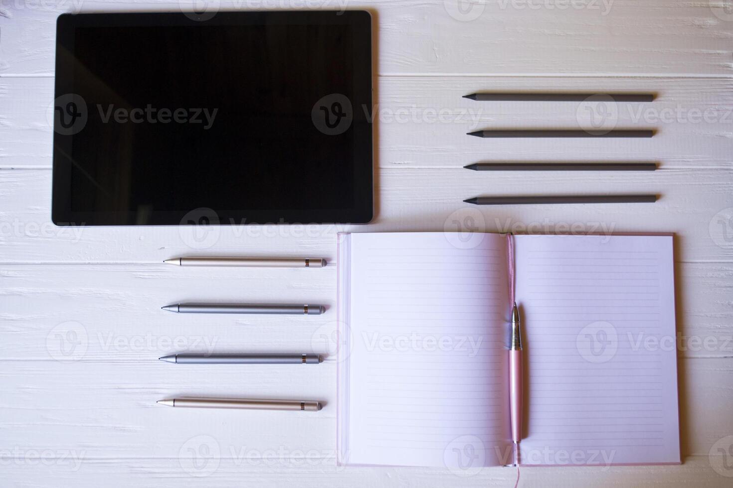 A tablet, an opened notepad, pencils and styluses on a white wooden desk. Top view. photo
