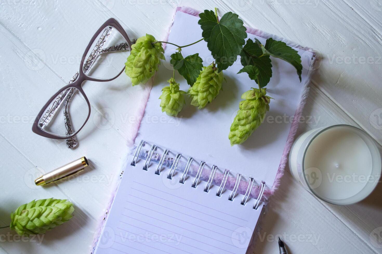 The opened notepad, pen, white candle, glasses and branches of hops as decoration on a white wooden table. Desktop still life with space for text. photo