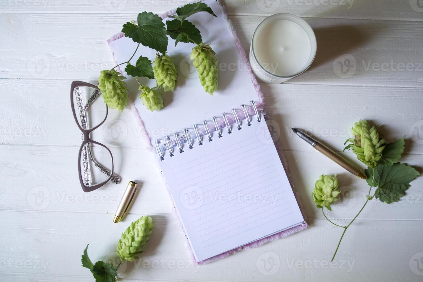 The opened notepad, pen, white candle, glasses and branches of hops as decoration on a white wooden table. Desktop still life with space for text. photo