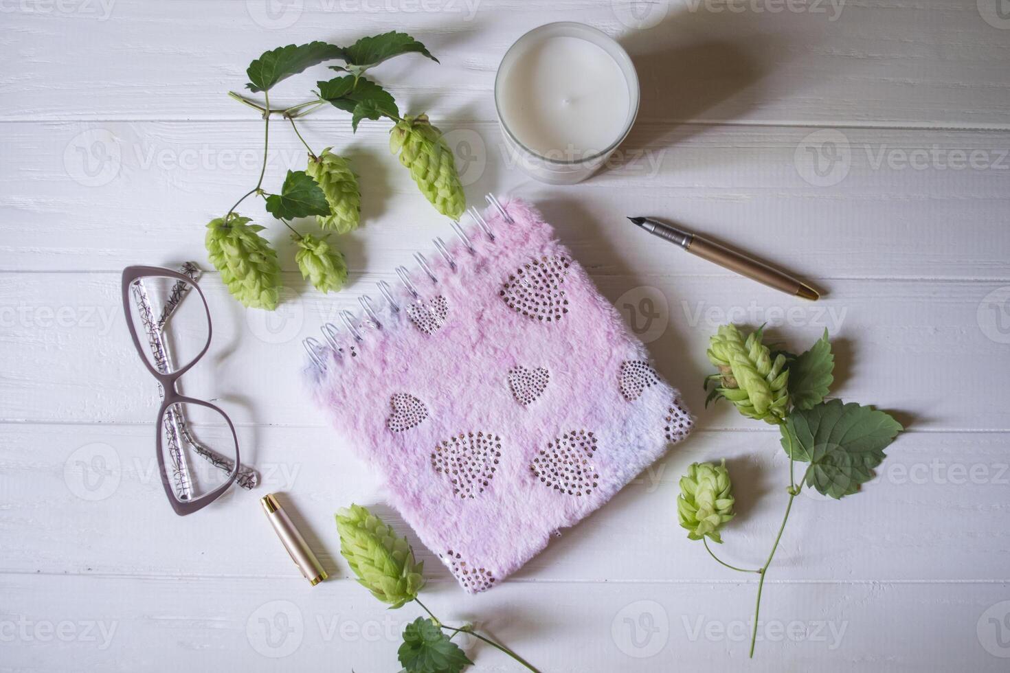 The closed pink notepad, pen, white candle and branches of hops as decoration on a white wooden table. Desktop still life. photo