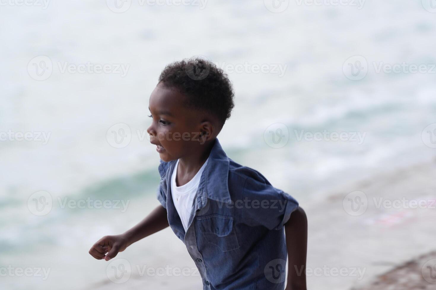 mezclado carrera africano y asiático chico es jugando a el al aire libre área. sonriente contento chico tiene divertido corriendo en el playa. retrato de chico estilo de vida con un único peinado. foto