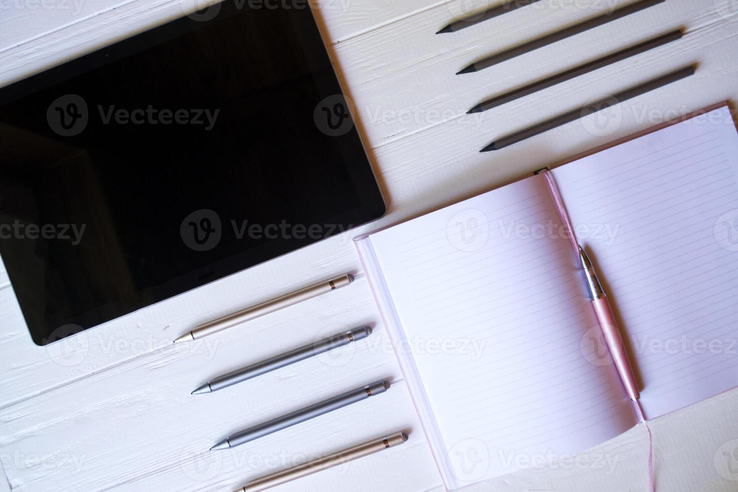 A tablet, an opened notepad, pencils and styluses on a white wooden desk. Top view. photo