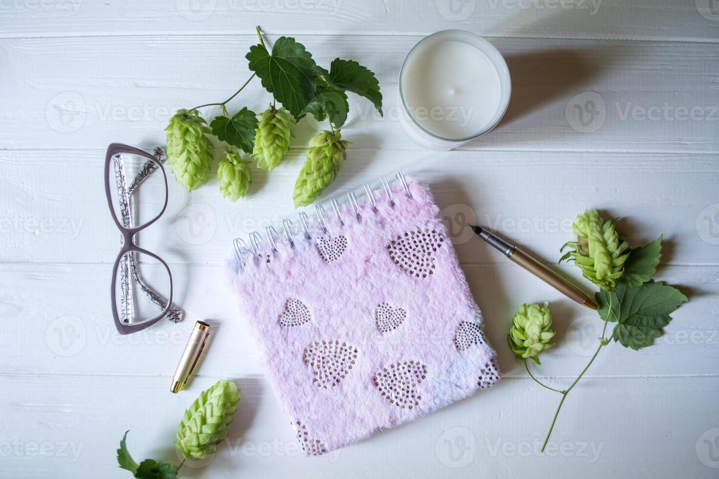The closed pink notepad, pen, white candle and branches of hops as decoration on a white wooden table. Desktop still life. photo