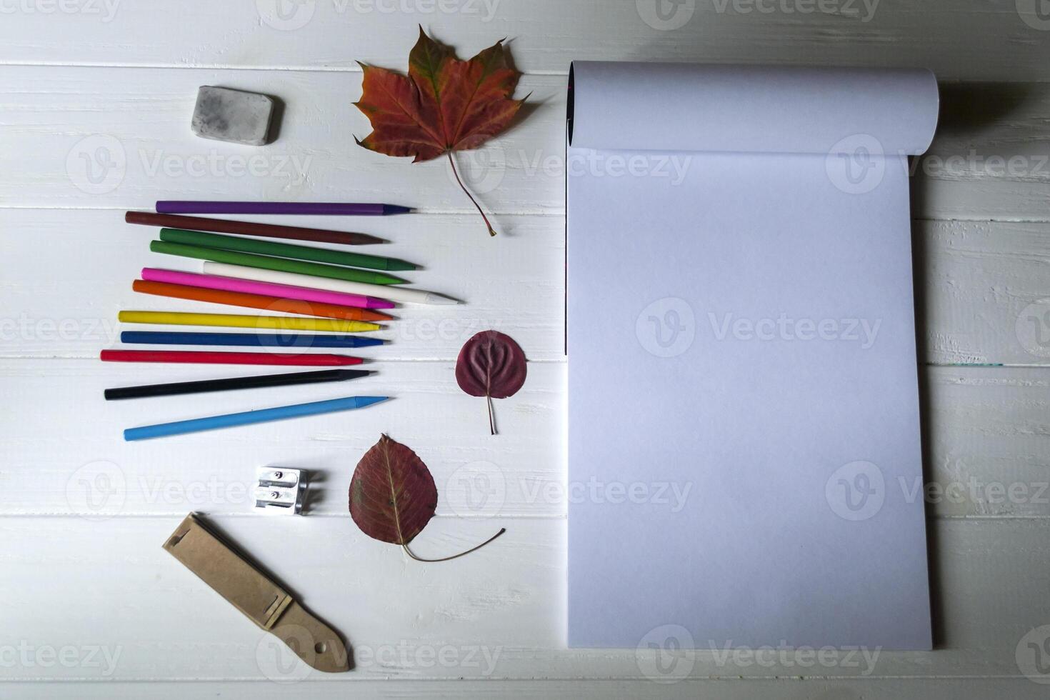 Empty sketch pad, colorful pencils, other equipment for drawing and fallen leaf on the white wooden desktop. Art background with place for text. photo