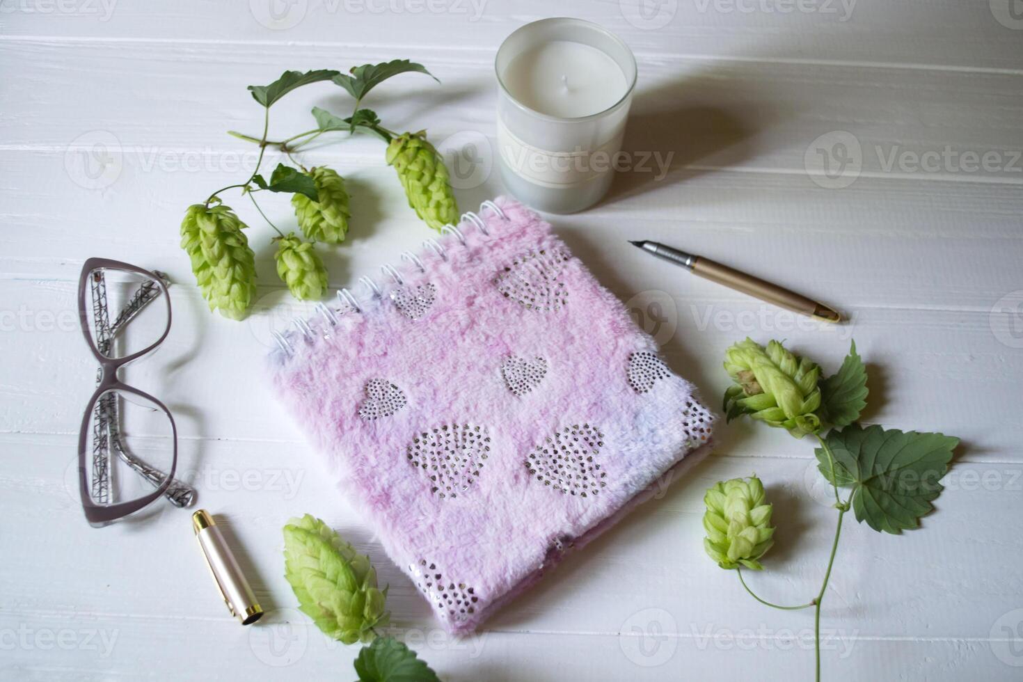 The closed pink notepad, pen, white candle and branches of hops as decoration on a white wooden table. Desktop still life. photo