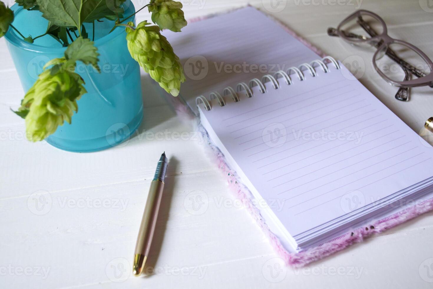 el abrió bloc, bolígrafo, blanco vela, lentes y ramas de lúpulo como decoración en un blanco de madera mesa. escritorio todavía vida con espacio para texto. foto