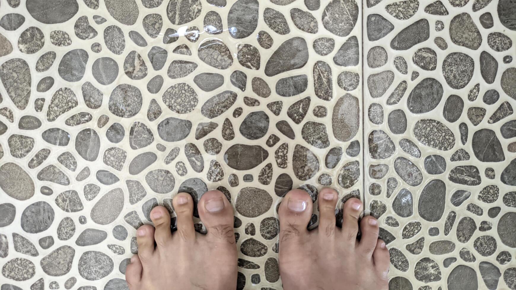 A bird's-eye view of tanned human feet standing on uniquely shaped ash tiles in a bathroom with water splashing and running down the drain. photo