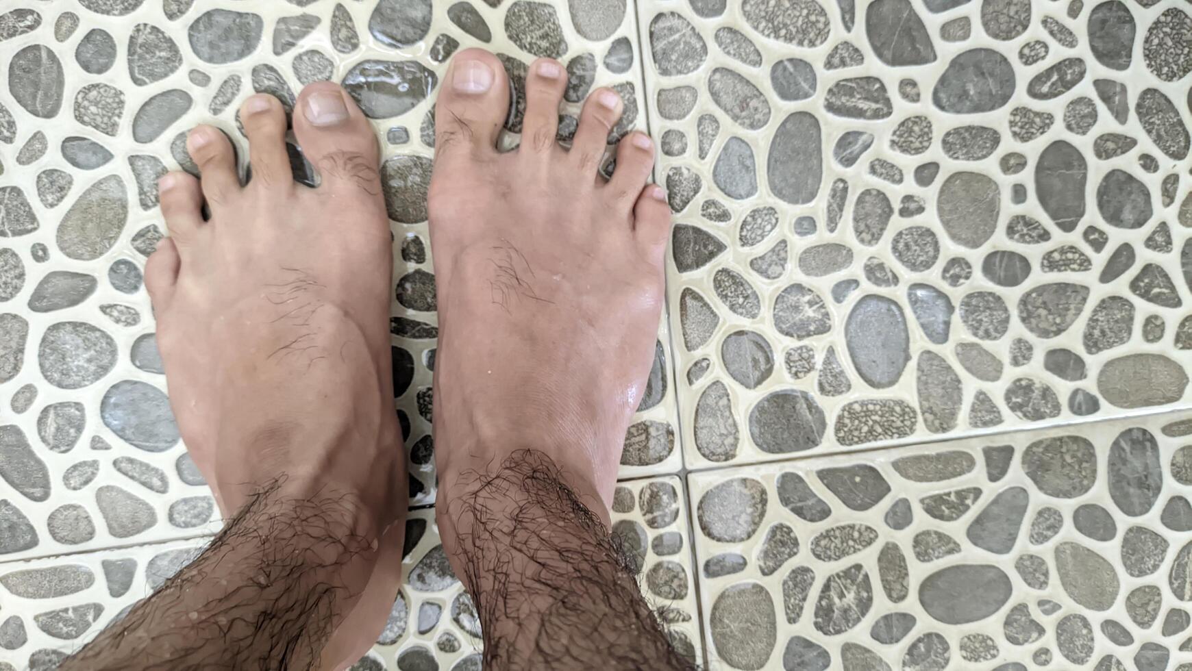 A bird's-eye view of tanned human feet standing on uniquely shaped ash tiles in a bathroom with water splashing and running down the drain. photo