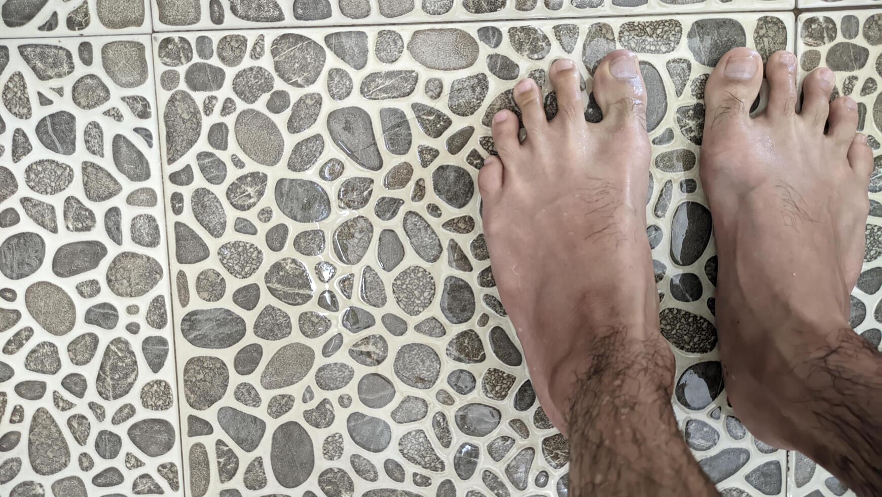 A bird's-eye view of tanned human feet standing on uniquely shaped ash tiles in a bathroom with water splashing and running down the drain. photo