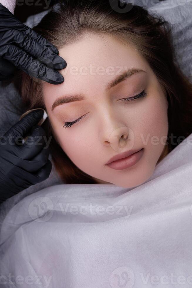 The master cleans the model's eyebrows from excess pigment after permanent makeup. PMU Procedure, Permanent Eyebrow Makeup. photo