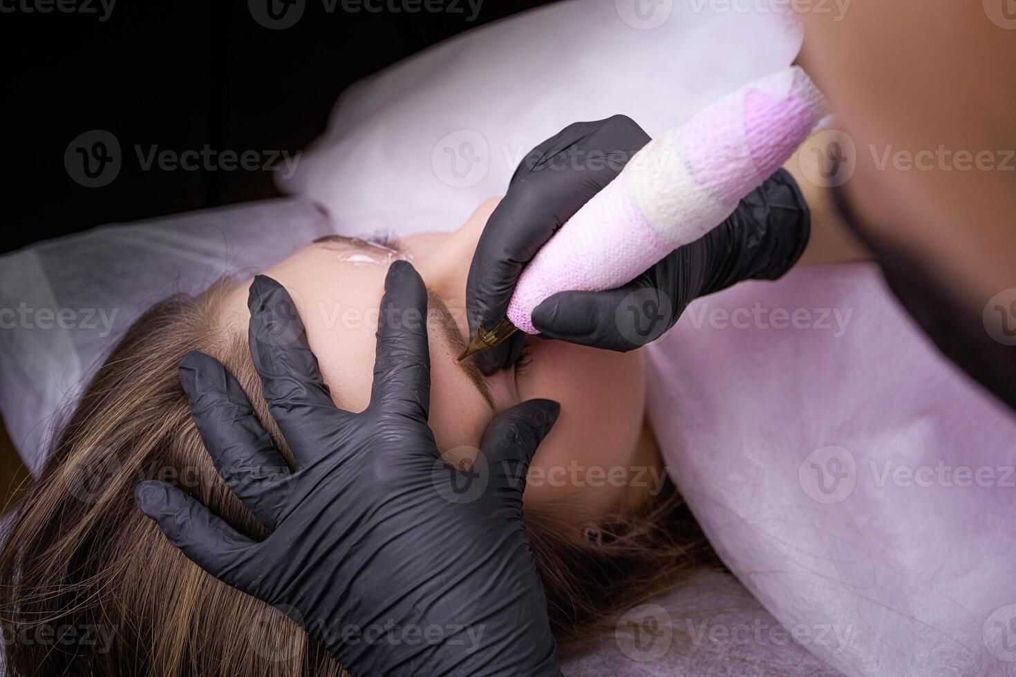 Applying permanent makeup on the eyebrows with a tattoo machine held by a master in black gloves. PMU Procedure, Permanent Eyebrow Makeup. photo
