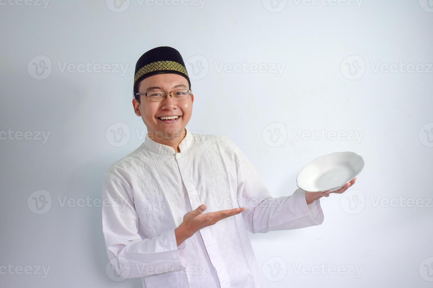 Asian Muslim man wearing glasses and white cloth pointing empty plate for fasting ramadhan. Isolated white background photo