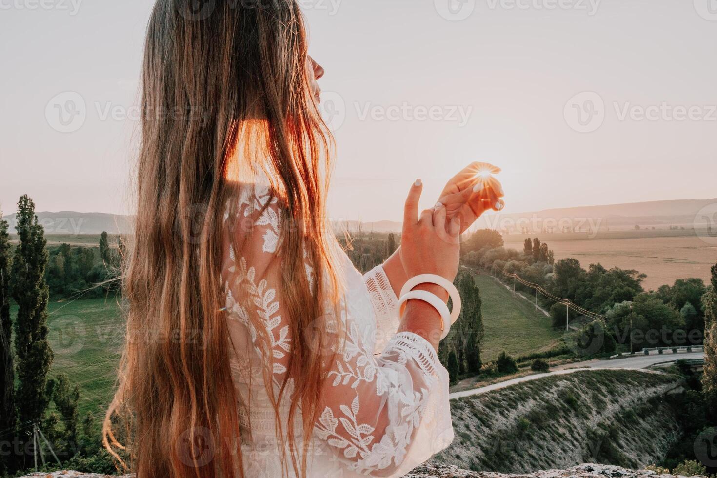 Happy woman in white boho dress on sunset in mountains. Romantic woman with long hair standing with her back on the sunset in nature in summer with open hands. Silhouette. Nature. Sunset. photo