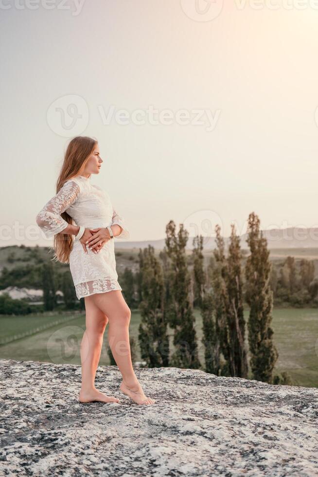 Happy woman in white boho dress on sunset in mountains. Romantic woman with long hair standing with her back on the sunset in nature in summer with open hands. Silhouette. Nature. Sunset. photo