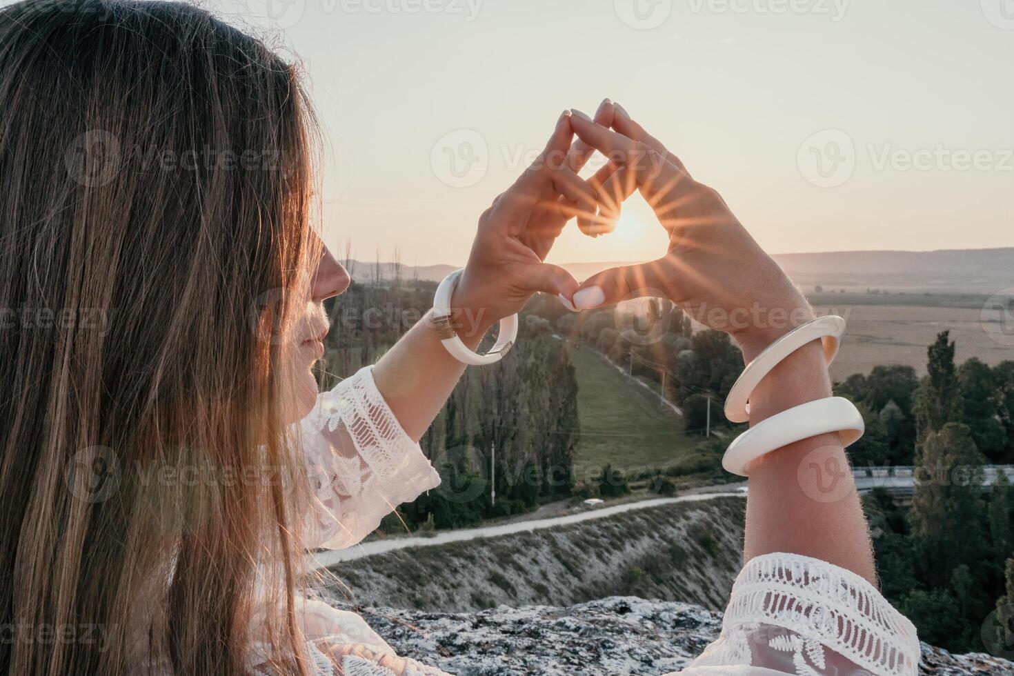 Happy woman in white boho dress making heart sign with hands on sunset in mountains. Romantic woman with long hair standing with her back on the sunset in nature in summer with open hands. photo