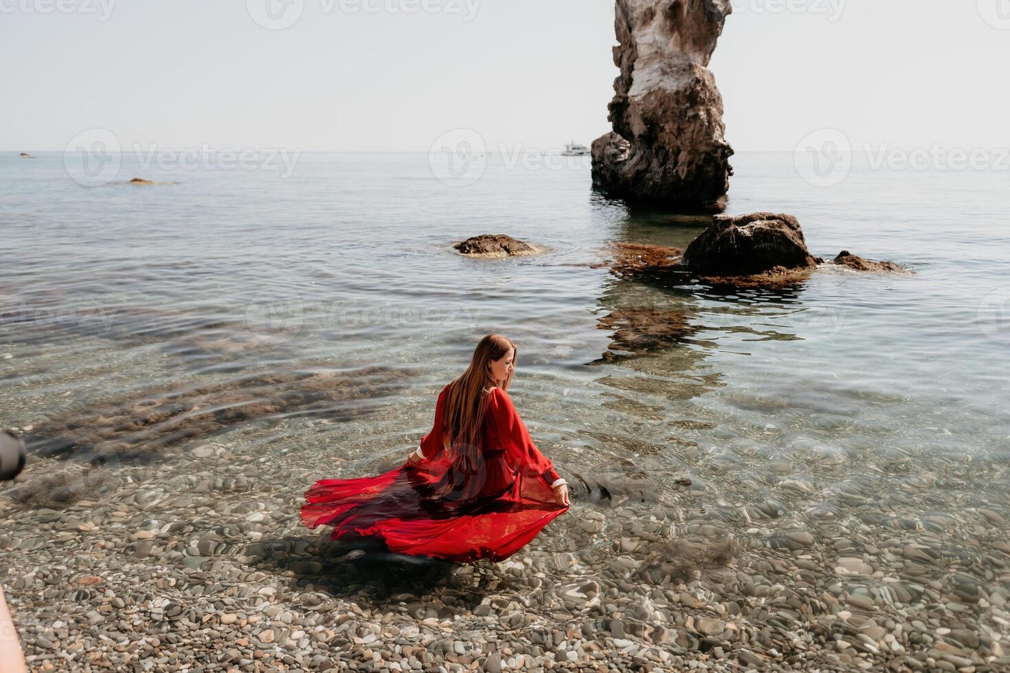 Woman travel sea. Happy tourist in red dress enjoy taking picture outdoors for memories. Woman traveler posing on the rock at sea bay surrounded by volcanic mountains, sharing travel adventure journey photo