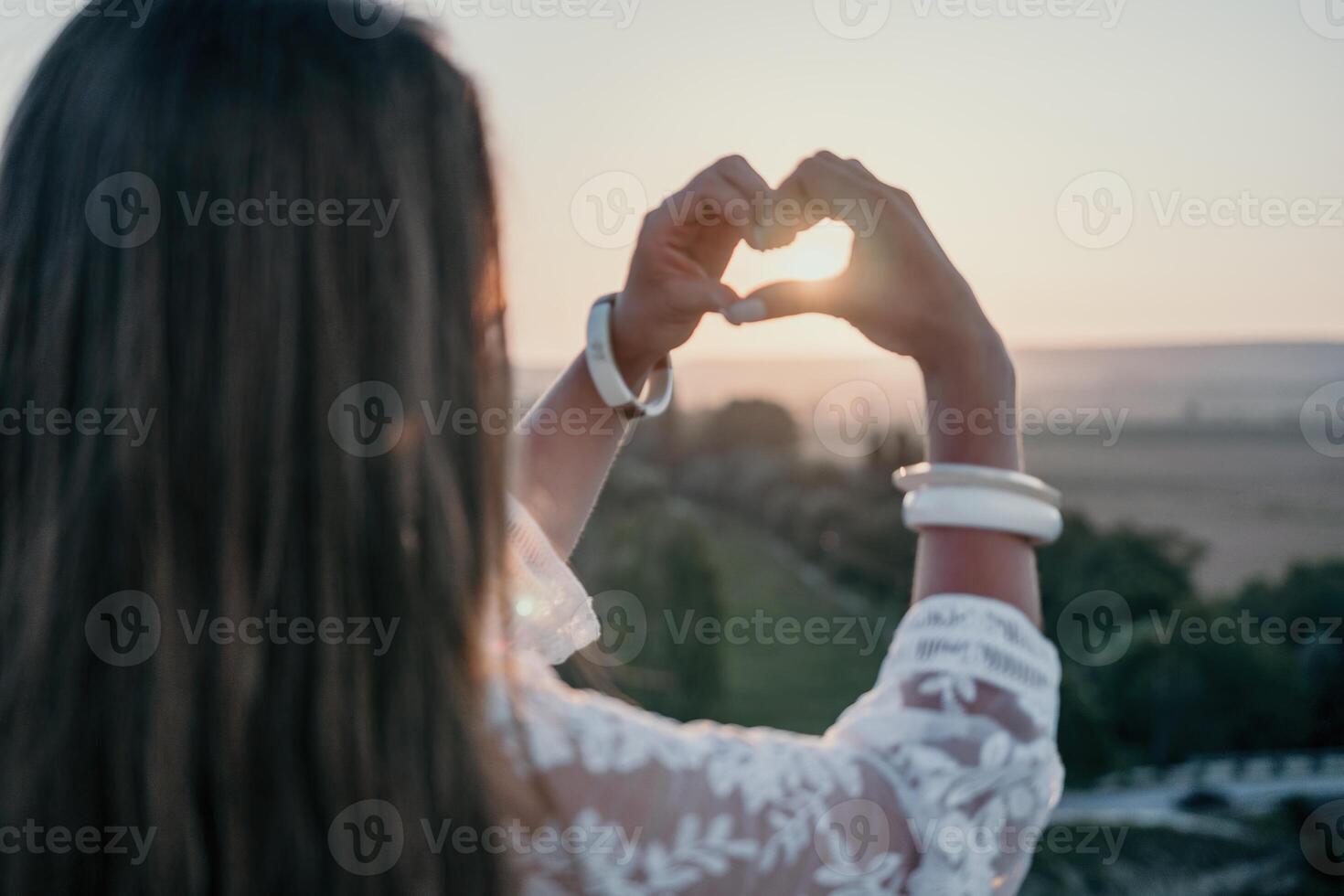 contento mujer en blanco boho vestir haciendo corazón firmar con manos en puesta de sol en montañas. romántico mujer con largo pelo en pie con su espalda en el puesta de sol en naturaleza en verano con abierto manos. foto