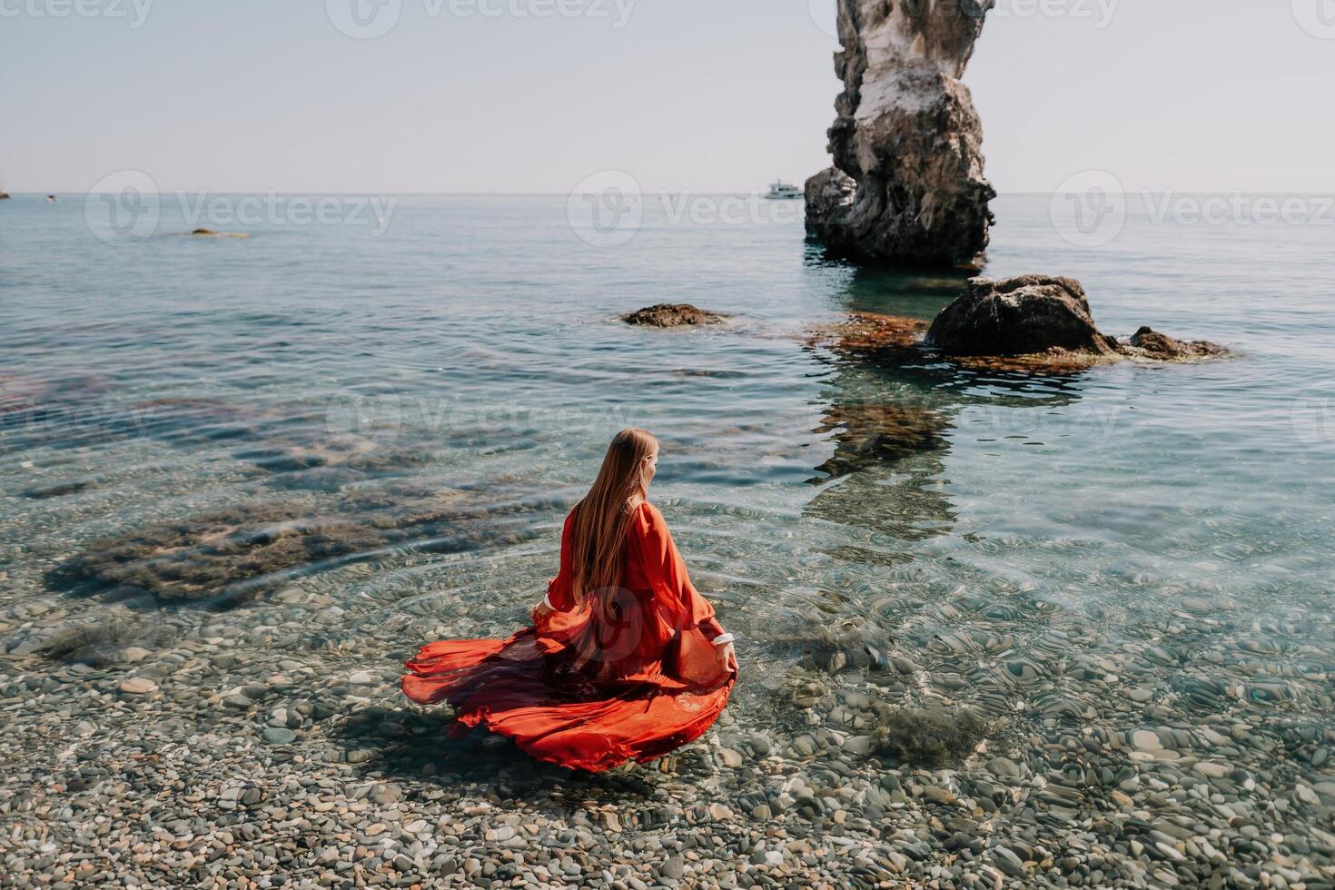 Woman travel sea. Happy tourist in red dress enjoy taking picture outdoors for memories. Woman traveler posing on the rock at sea bay surrounded by volcanic mountains, sharing travel adventure journey photo