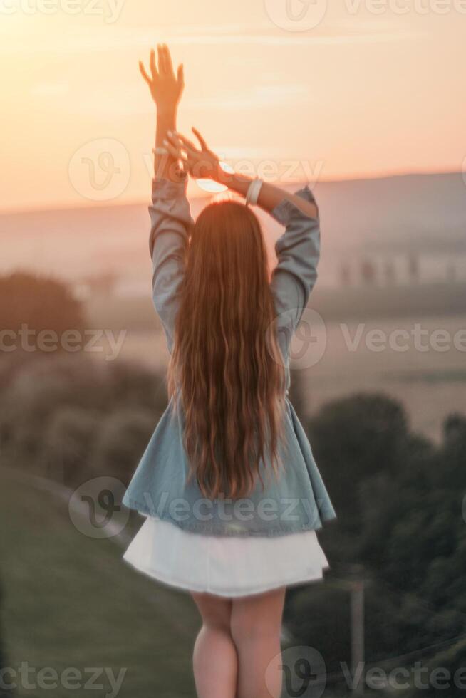 Happy woman standing with her back on the sunset in nature in summer with open hands. Romantic beautiful bride in white boho dress posing with mountains on sunset photo