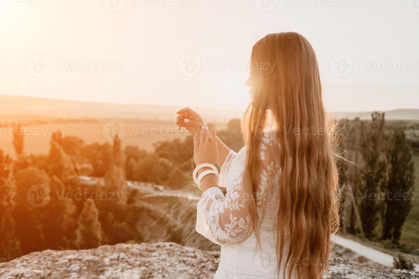 Happy woman in white boho dress on sunset in mountains. Romantic woman with long hair standing with her back on the sunset in nature in summer with open hands. Silhouette. Nature. Sunset. photo