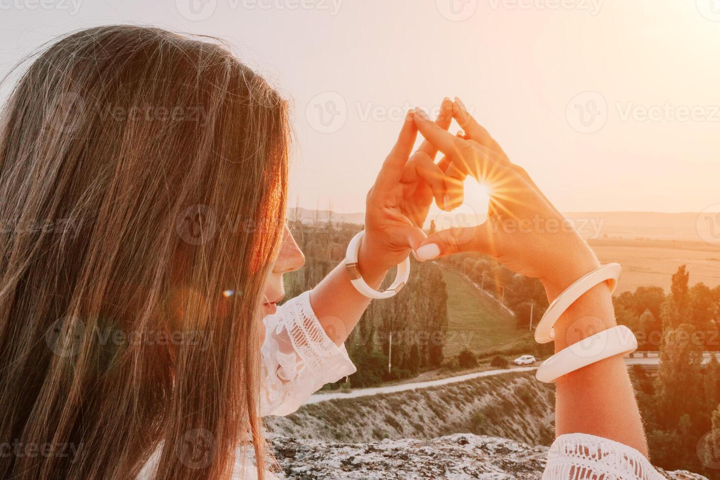 Happy woman in white boho dress making heart sign with hands on sunset in mountains. Romantic woman with long hair standing with her back on the sunset in nature in summer with open hands. photo