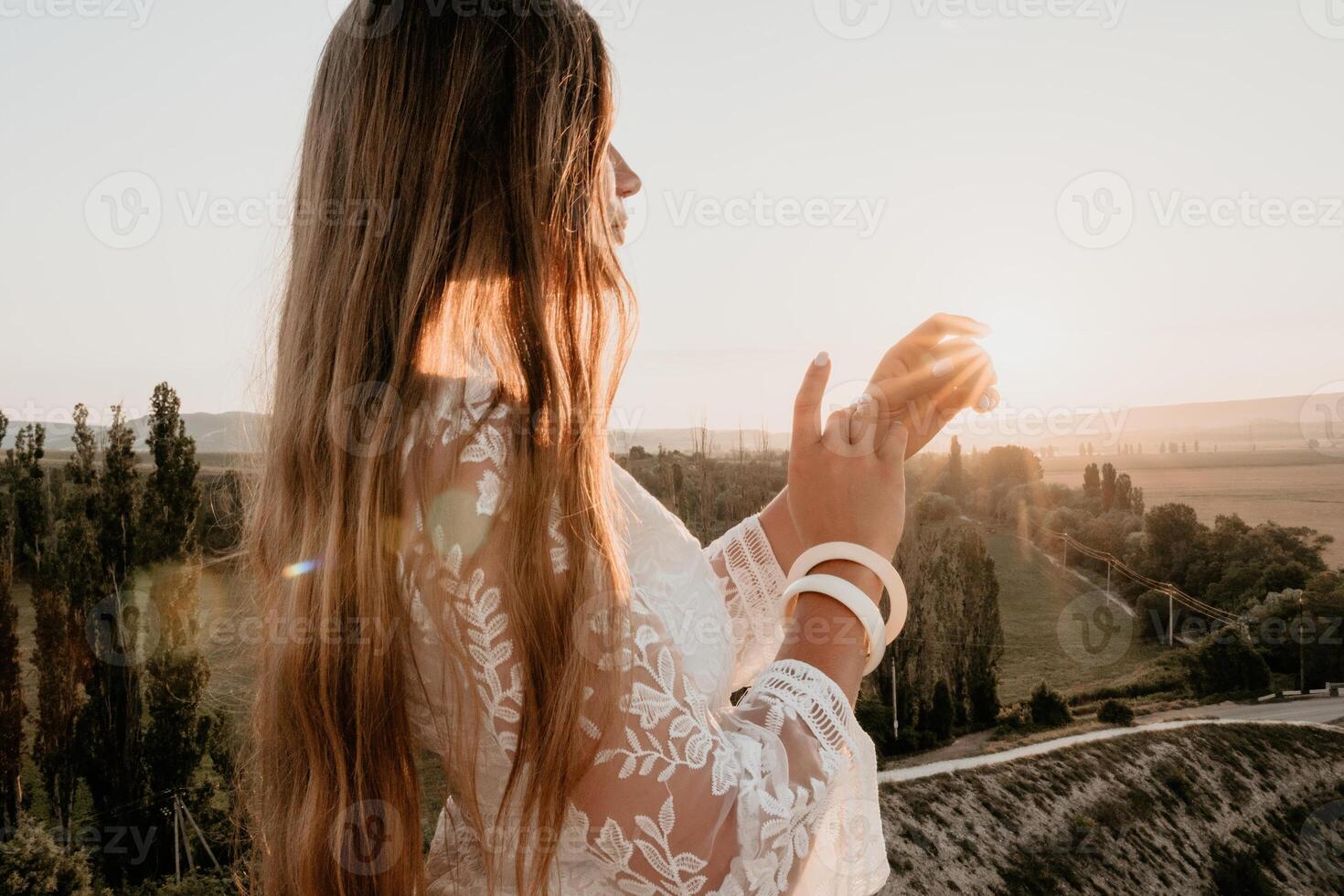 Happy woman in white boho dress on sunset in mountains. Romantic woman with long hair standing with her back on the sunset in nature in summer with open hands. Silhouette. Nature. Sunset. photo