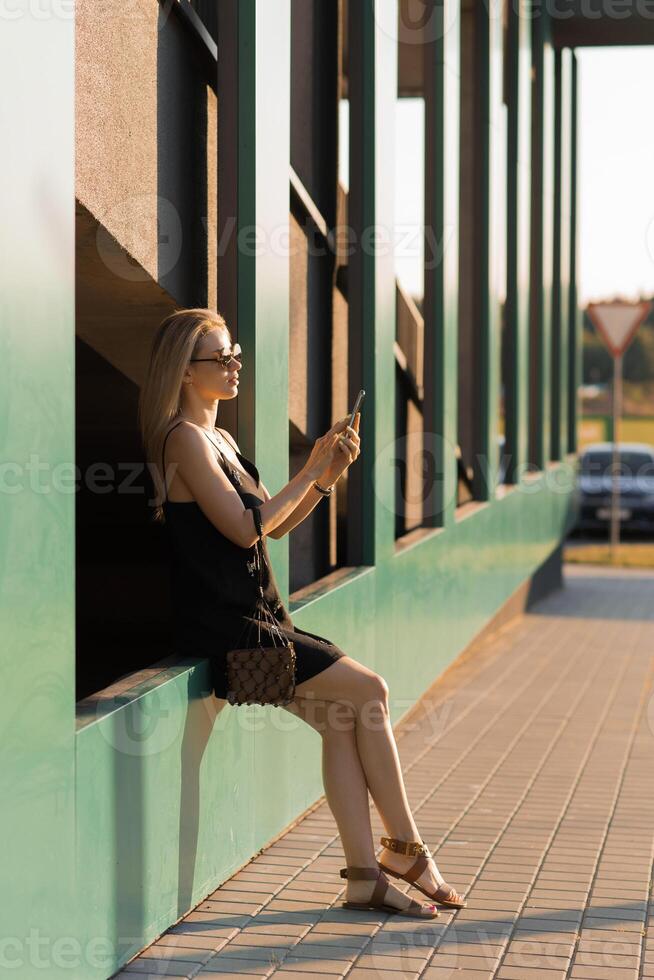 Young cheerful stylish woman using cell phone and texting message on city street. Beautiful happy hipster girl holding smart phone and surfing internet outdoors. Online communication photo